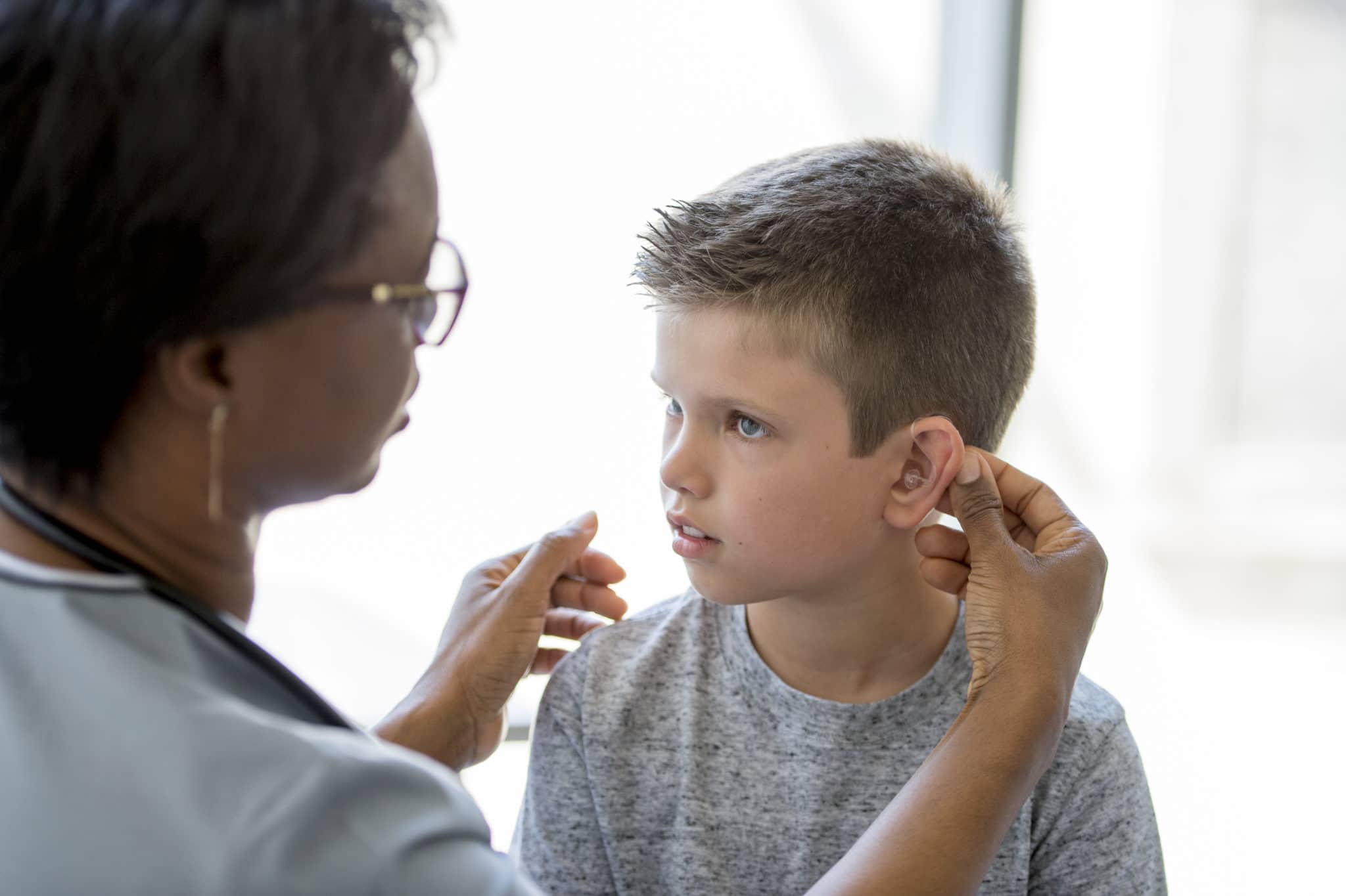 A little boy is sitting on an examining table in a doctors office - a healthcare professional is implanting a hearing aid for the child.