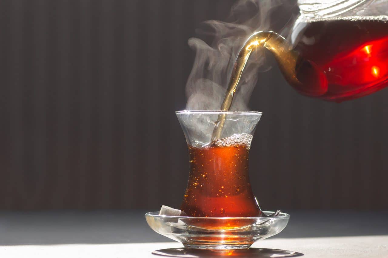 Glass cup of brewed black turkish tea pouring from teapot, traditional hot drink concept