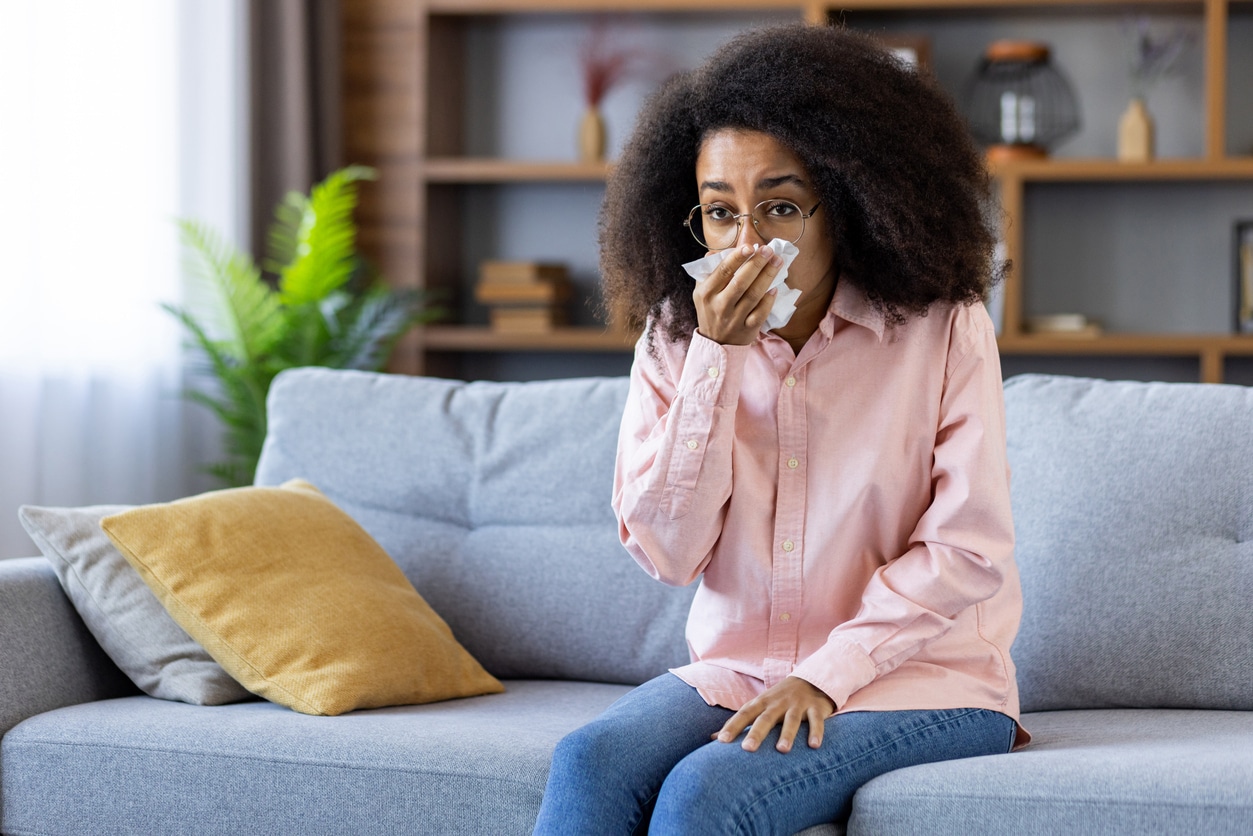 Woman sneezing into tissue, sitting on sofa at home during cold flu season.