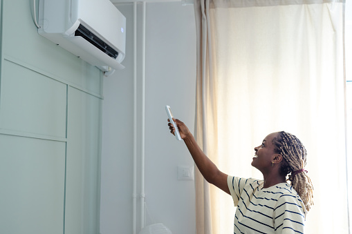 A smiling woman uses a remote to adjust a wall-mounted air conditioner in a bright bedroom